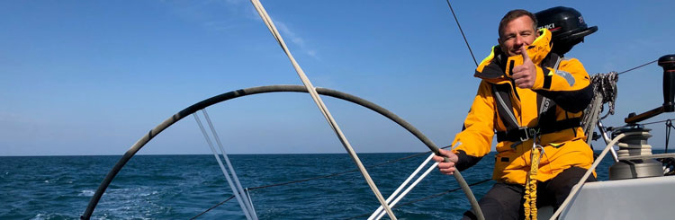 Foto a bordo di una barca a vela di una persona con una giacca arancione che tiene il timone e guarda la telecamera con un pollice in alto